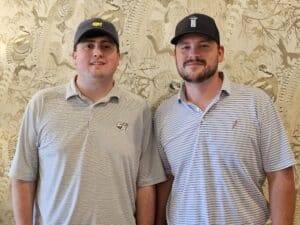 Two men wearing striped polo shirts and baseball caps stand next to each other before patterned wallpaper, both smiling softly at the camera.
