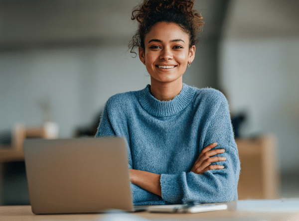 A woman with curly hair in a blue sweater sits at a desk, arms crossed, smiling. An open laptop and notebook are in front of her.
