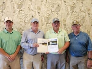 Four men in golf attire stand before a patterned wall; two center men display an open folder with papers, all smiling at the camera.