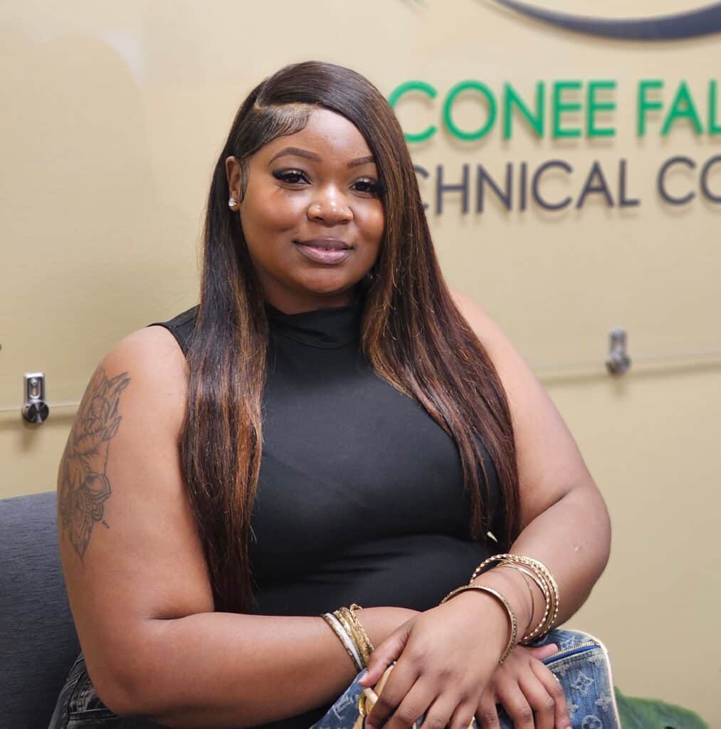 A woman with long straight hair and a tattoo on her upper arm smiles at the camera, wearing a sleeveless black top and bracelets; a Technical College sign is in the background.