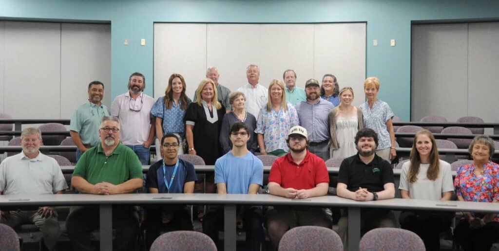 A group of 22 diverse people pose together in a classroom with gray tables and a blue-teal wall, arranged in seated and standing rows.