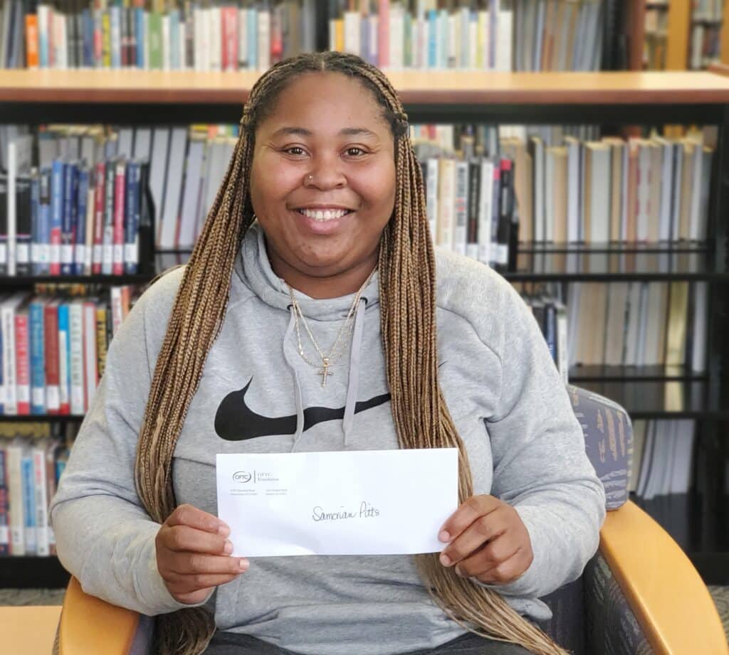 A smiling person with long braids, in a gray Nike hoodie, sits in a library holding an envelope labeled Samantha Pitts. Bookshelves are behind.