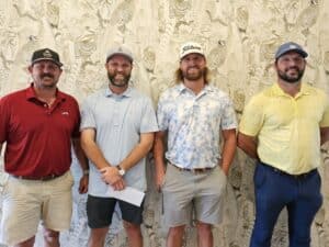 Four men stand together in front of a patterned wall, all wearing golf shirts, shorts or pants, caps, and smiling at the camera.