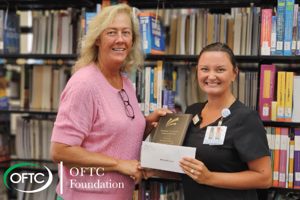 Two women are smiling; one is wearing scrubs and holding a plaque and an envelope. They are standing in front of library shelves, with the OFTC Foundation logo visible in the bottom left corner.