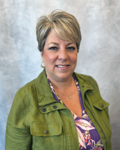Woman with short blonde hair, dressed in a green jacket layered over a floral top, gold hoop earrings, and a cross necklace, smiling while standing against a light gray textured background.