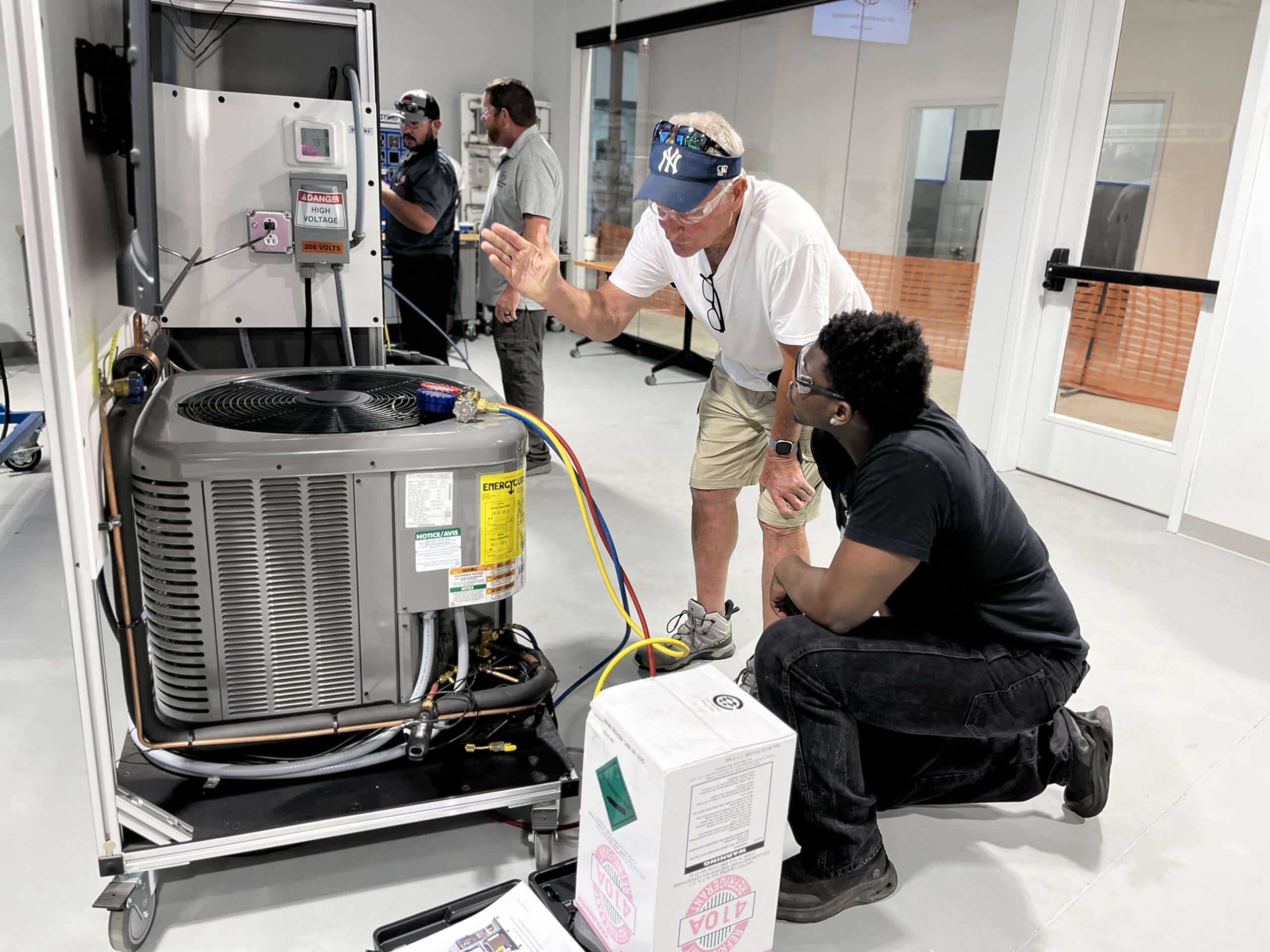 Two men examine and discuss an HVAC unit in a brightly lit room with visible equipment and tools. One man gestures toward the unit while the other kneels, listening carefully. Additional people are working in the background.
