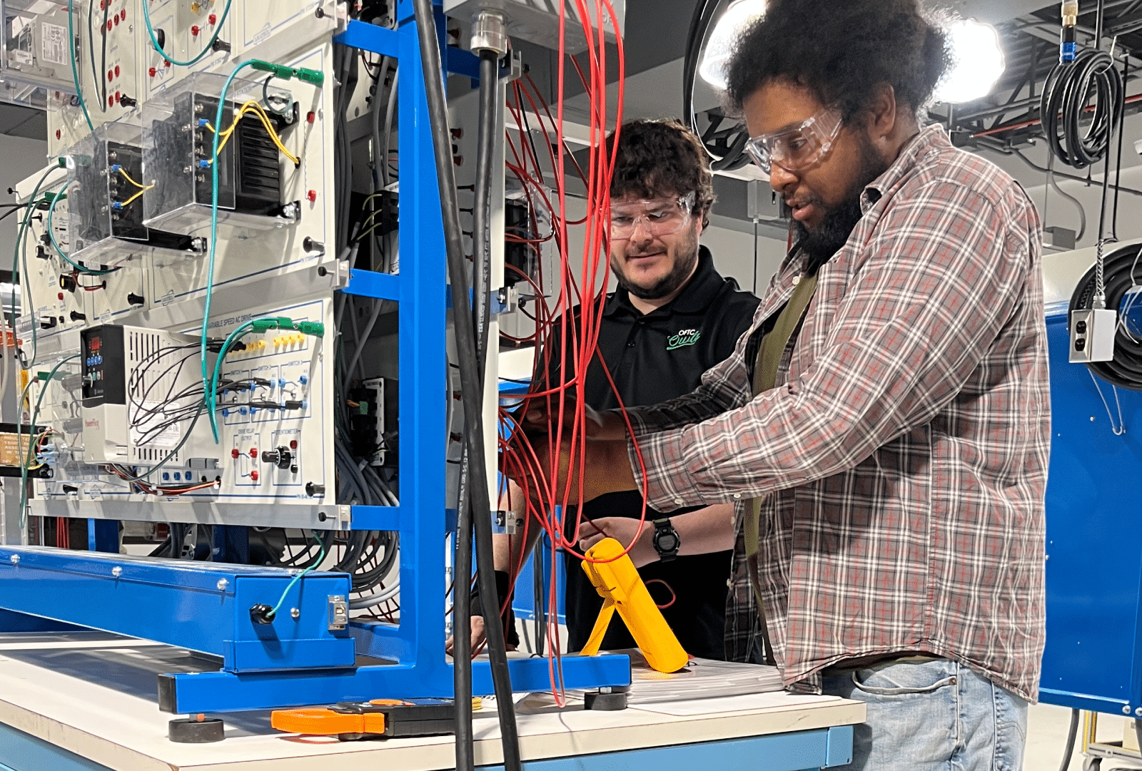 Two men in safety glasses handle electrical wiring and equipment in a laboratory, with cables and technical devices scattered around their blue and white workstation.