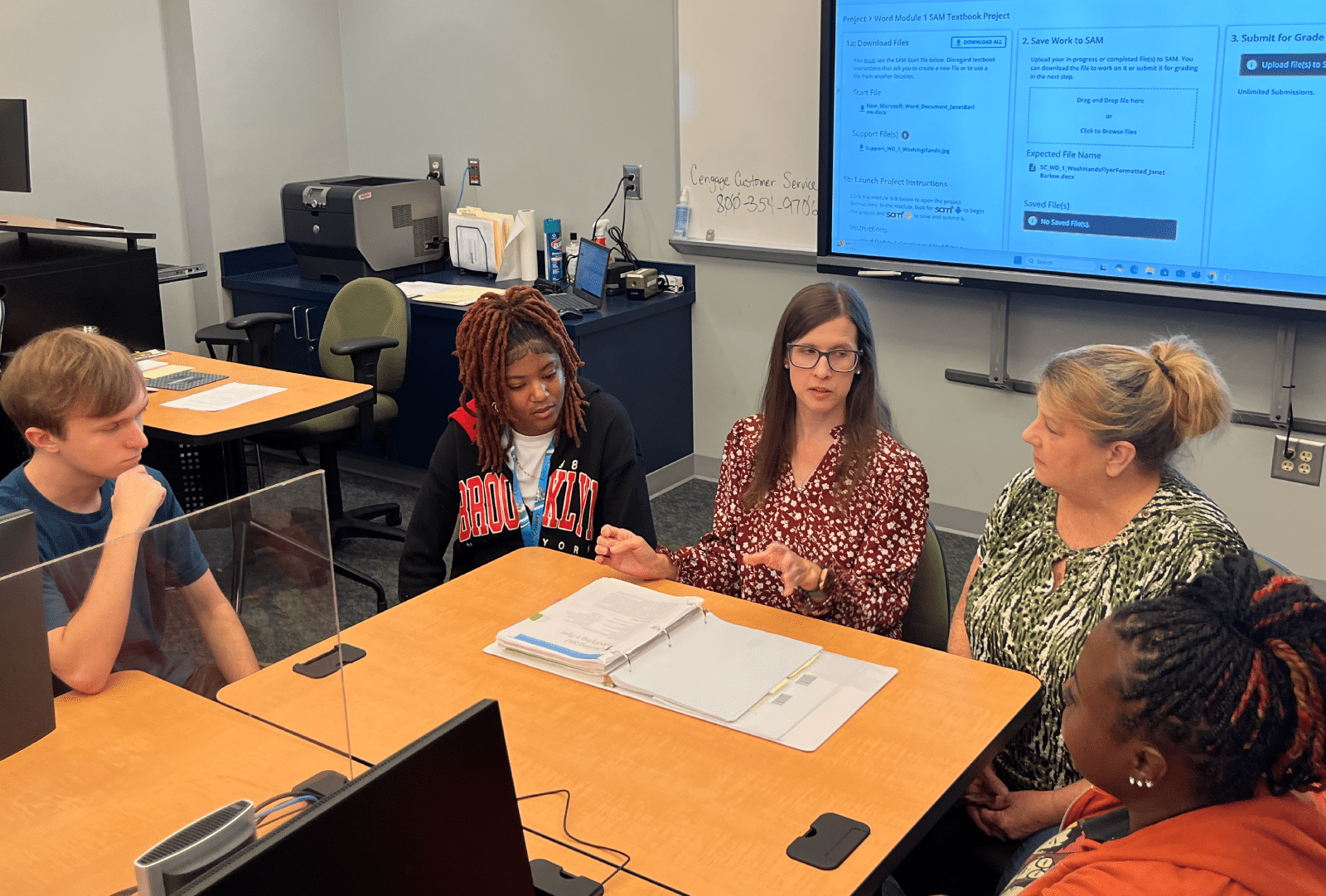 Five people are seated at a table in a classroom, engaged in discussion over documents. Behind them, a computer screen displays a website. One individual gestures as they speak while the others listen attentively.