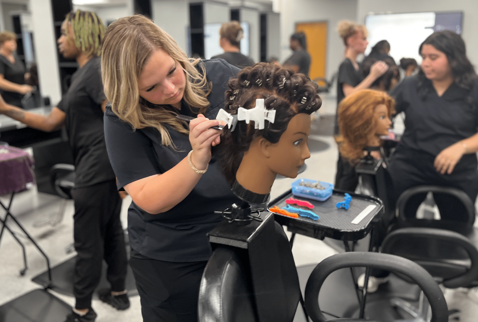A woman is styling a mannequin head with curling clips in a salon classroom, while other students nearby are also working on mannequin heads.