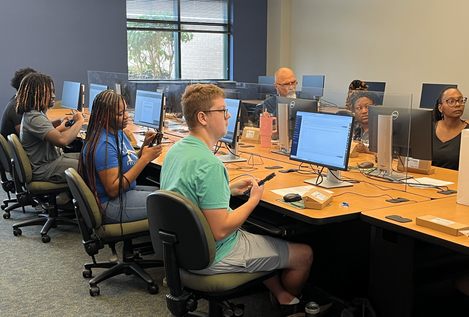 A group of people sits at computer stations in an office or classroom, each working on desktop computers separated by clear dividers. Some individuals are typing, while others focus on their screens or engage in conversation.