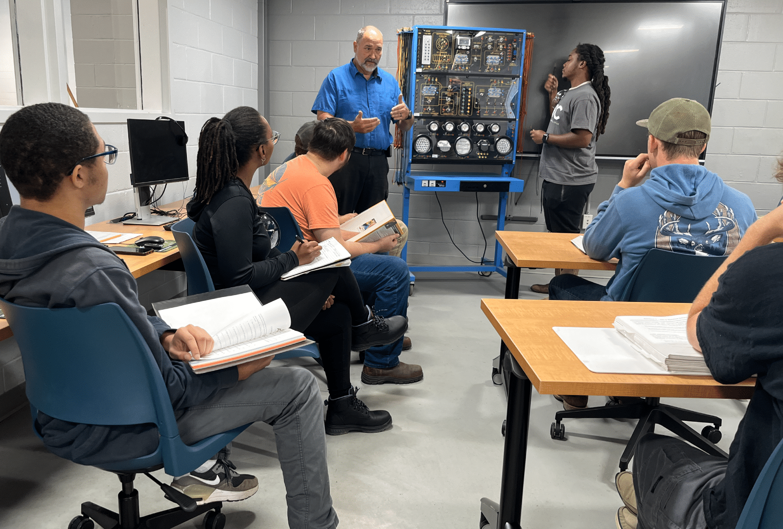 A teacher stands by a control panel, instructing five students who are seated at desks with open books in a classroom. One student stands near the teacher, listening attentively.