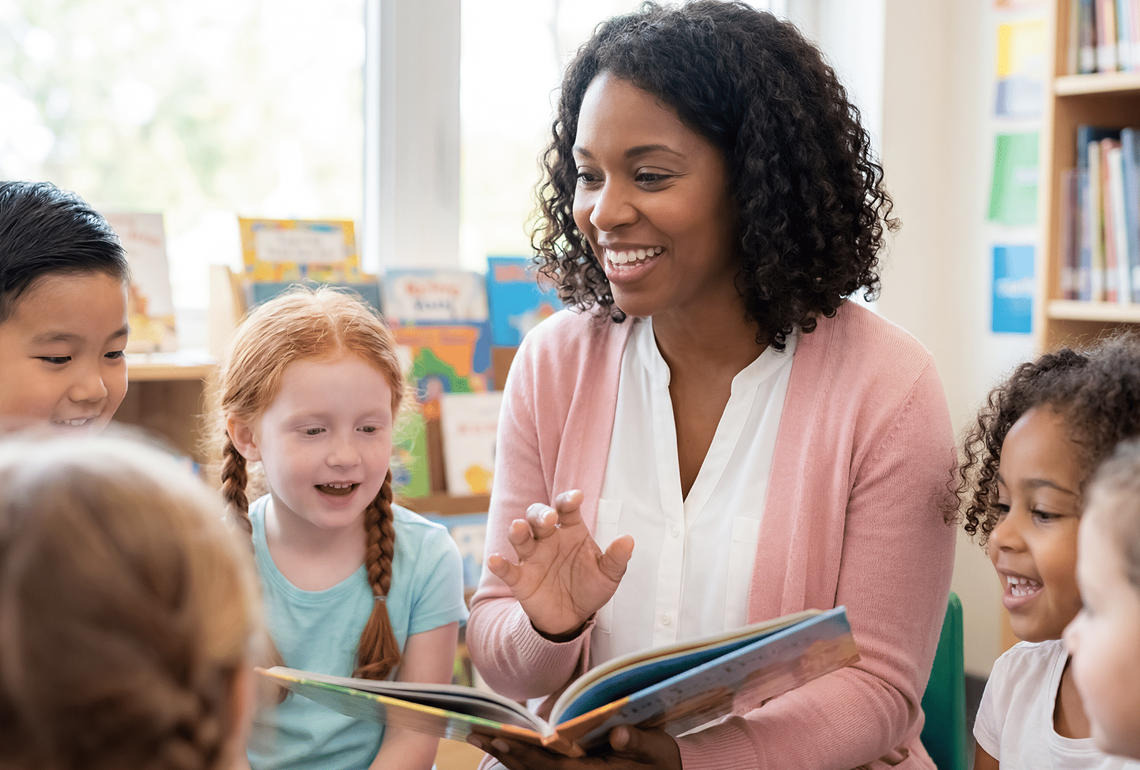 A cheerful woman reads a book to several young children in a well-lit classroom, surrounded by bookshelves and large windows in the background.