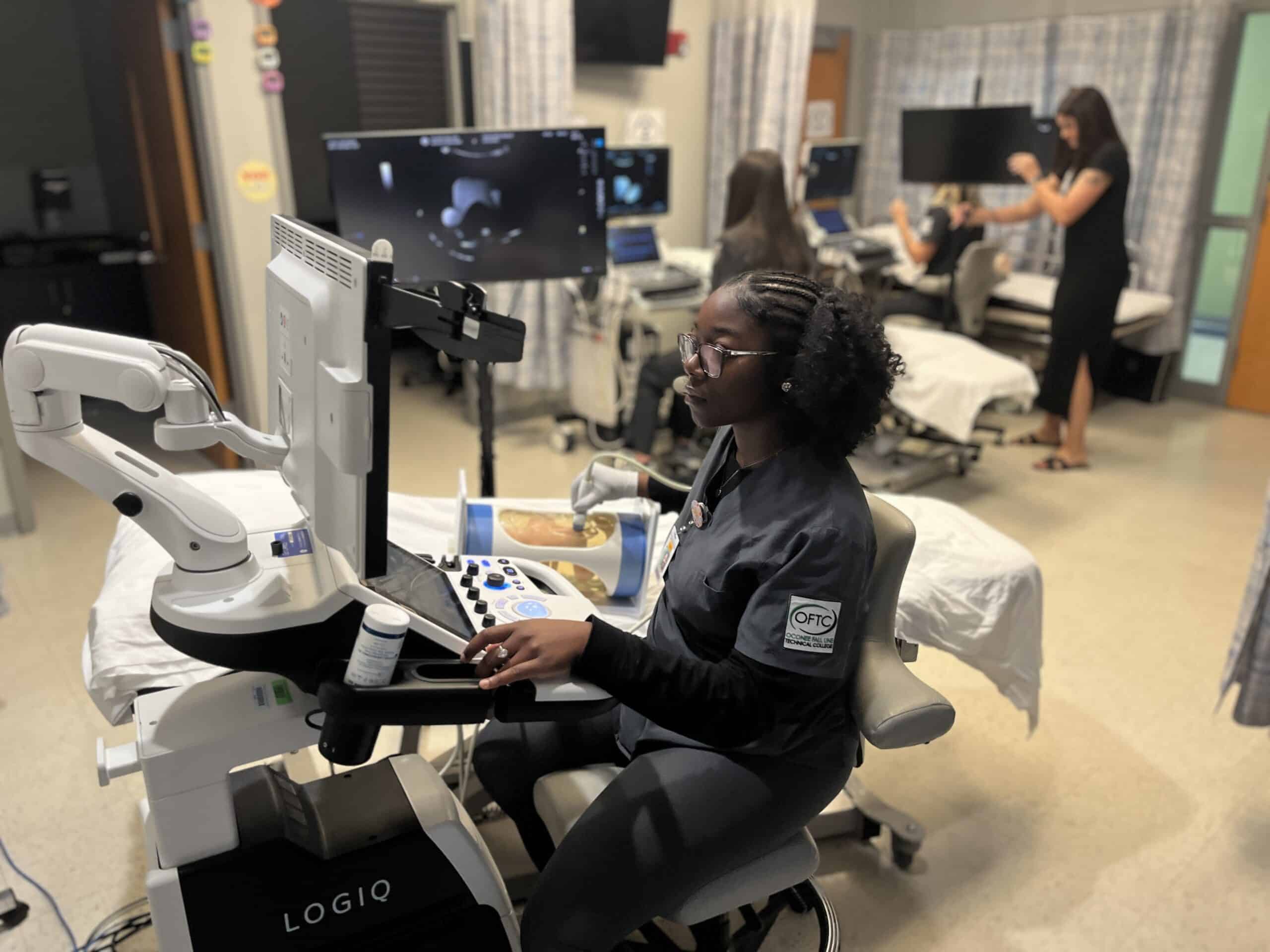 A woman uses a medical ultrasound machine in a clinical training room, with other individuals working at computers and ultrasound stations in the background.