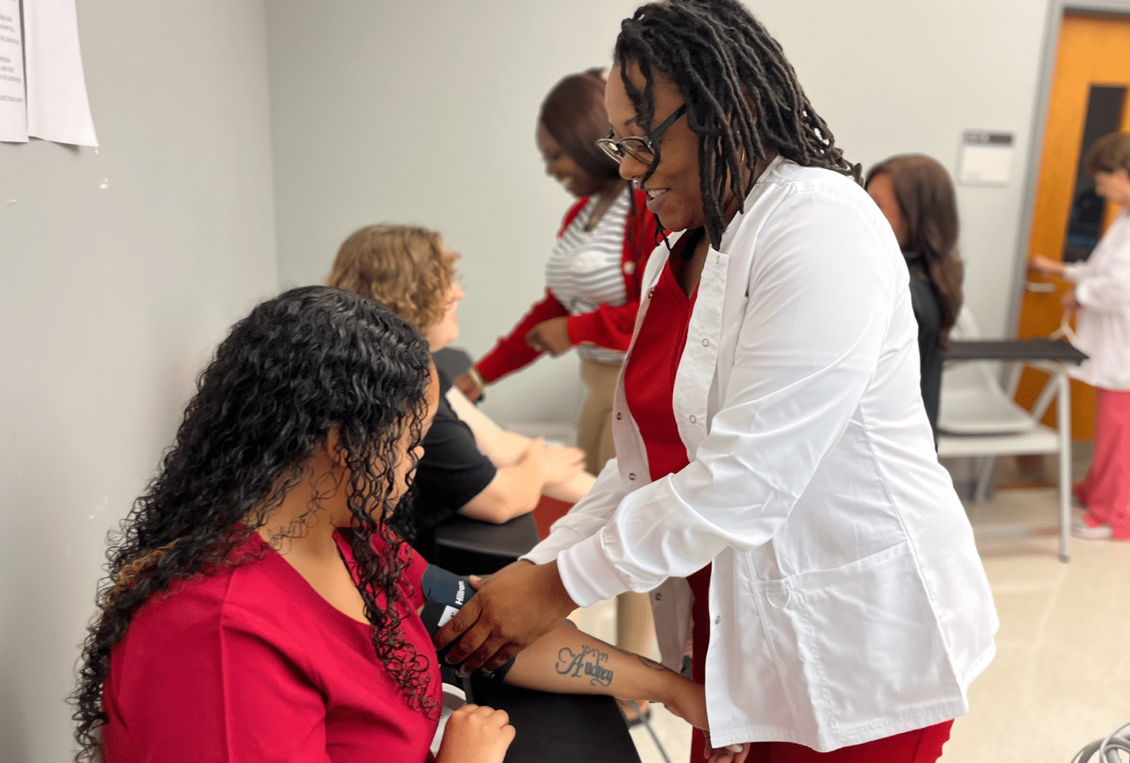 A healthcare worker takes a patient’s blood pressure in a clinic setting. Other staff and patients are visible in the background, all engaged in similar activities.