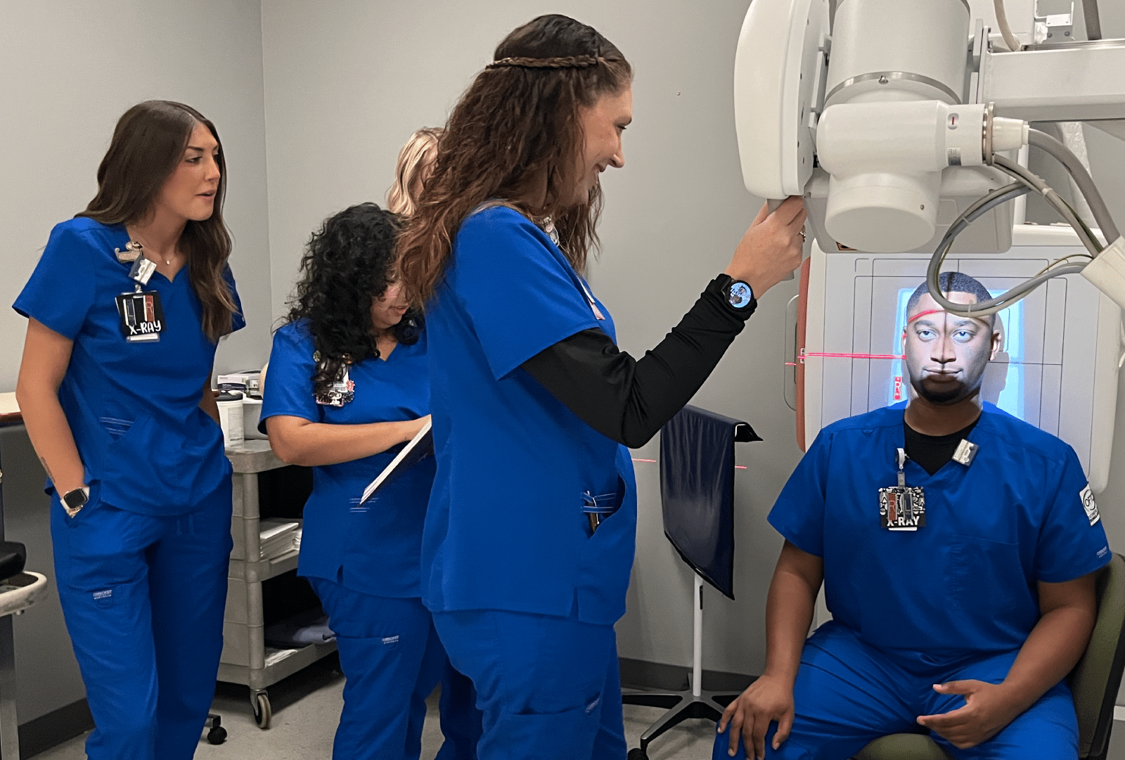 Four medical professionals in blue scrubs prepare a patient for a facial X-ray in a clinical setting; one operates the machine while others observe and take notes.