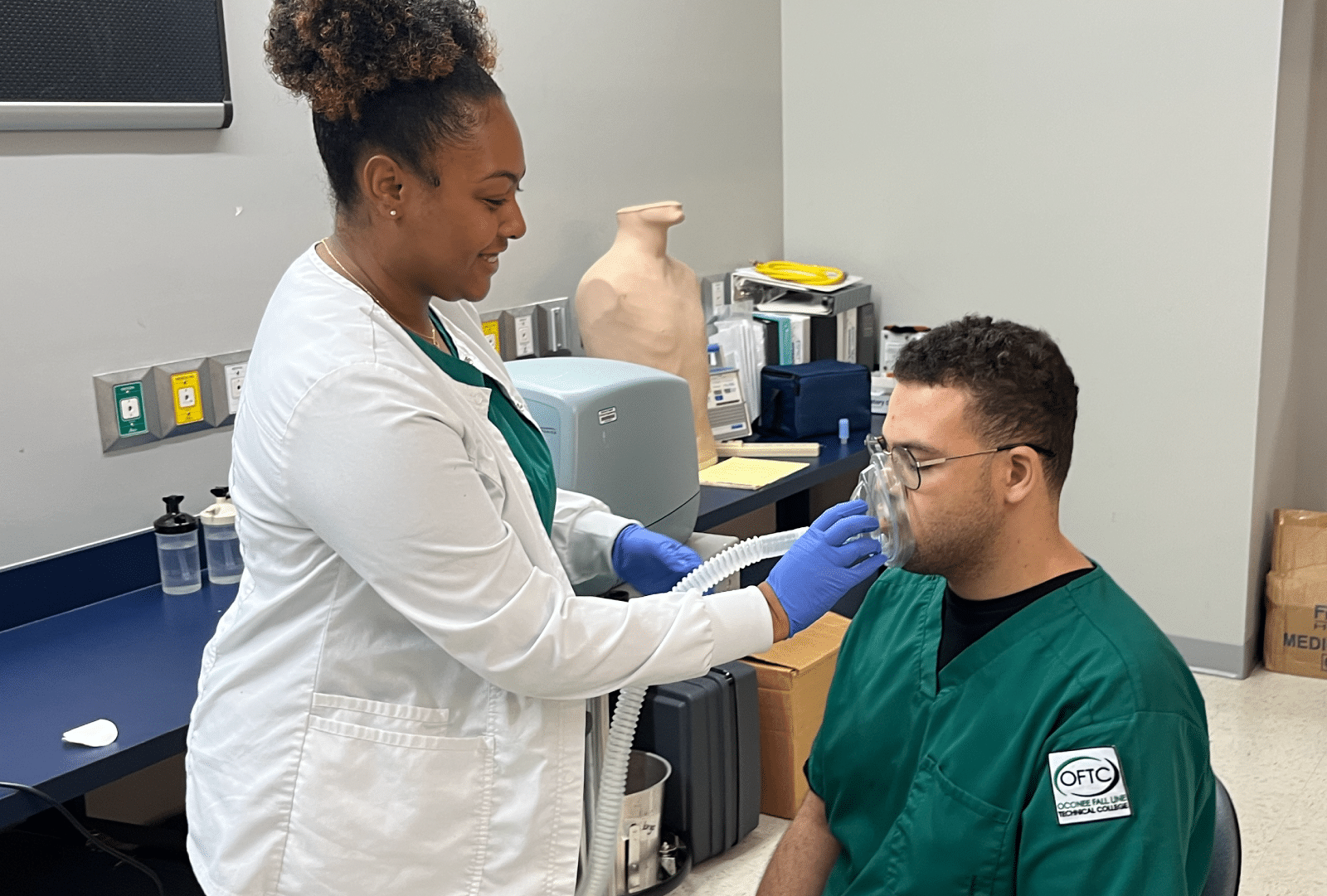 A healthcare worker wearing a white coat helps a seated individual in green scrubs who is using an oxygen mask, inside a medical classroom. Medical equipment and supplies can be seen in the background.