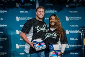 Two people smile while holding boxed iPads with blue ribbons. They wear matching black Owls OFTC jerseys and stand in front of a blue Technical College System of Georgia backdrop.
