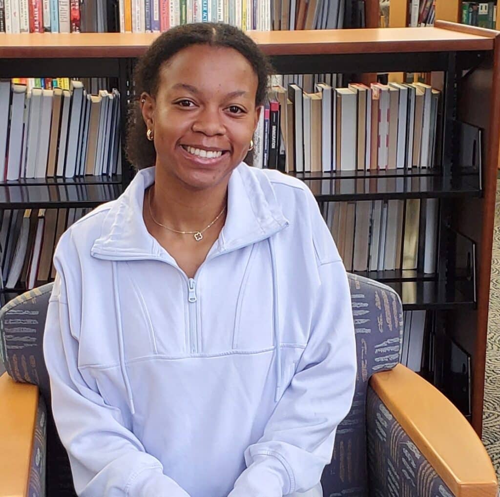 A woman smiling while seated in a library, dressed in a light purple zip-up jacket. Bookshelves lined with books can be seen behind her.