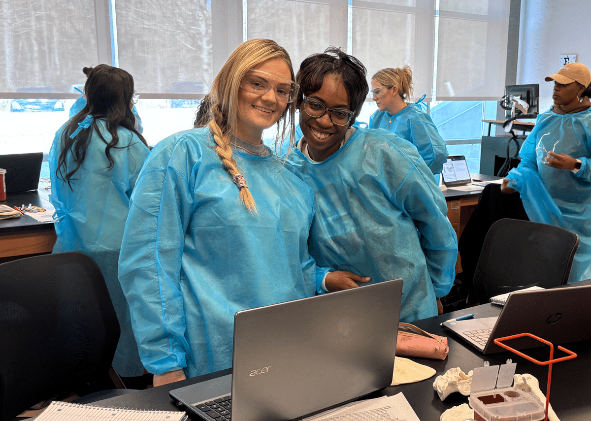 Two smiling OFTC students in blue lab gowns and safety glasses smile at a desk with a laptop in a bright science lab classroom. In the background, other students dressed similarly work with lab equipment.