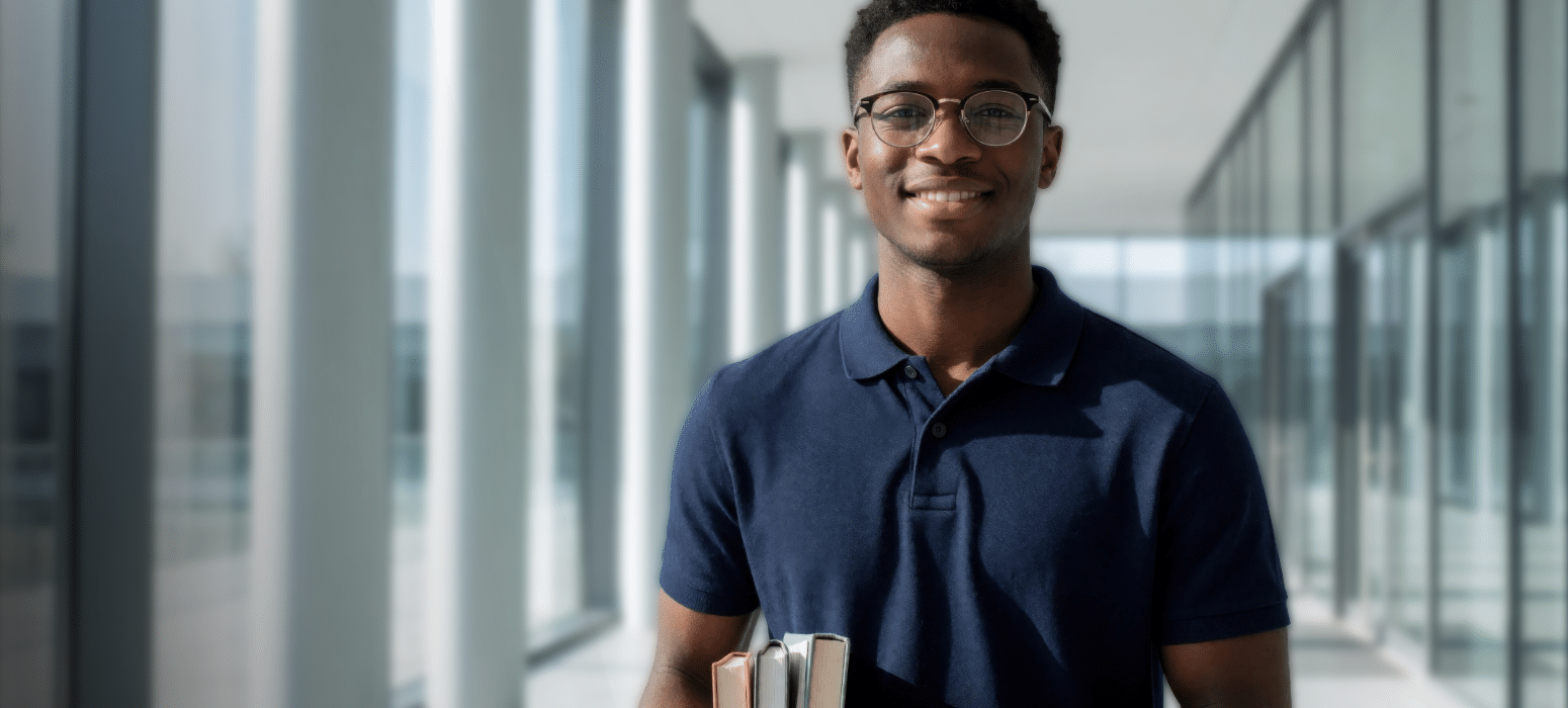 A smiling young man in glasses and a navy blue polo shirt stands in a bright hallway with large windows, holding several books.