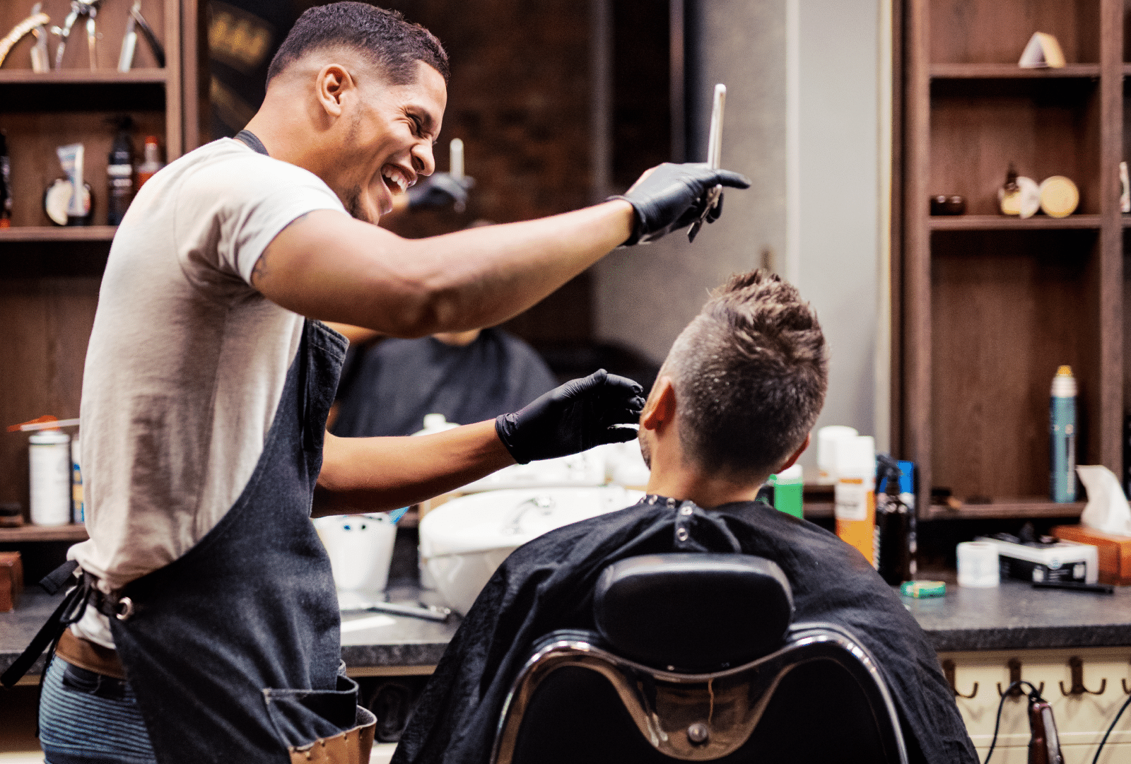 A smiling barber wearing gloves and an apron cuts a customer’s hair in a modern barbershop, using scissors while the client sits in a chair facing away. Shelves with grooming products are in the background.