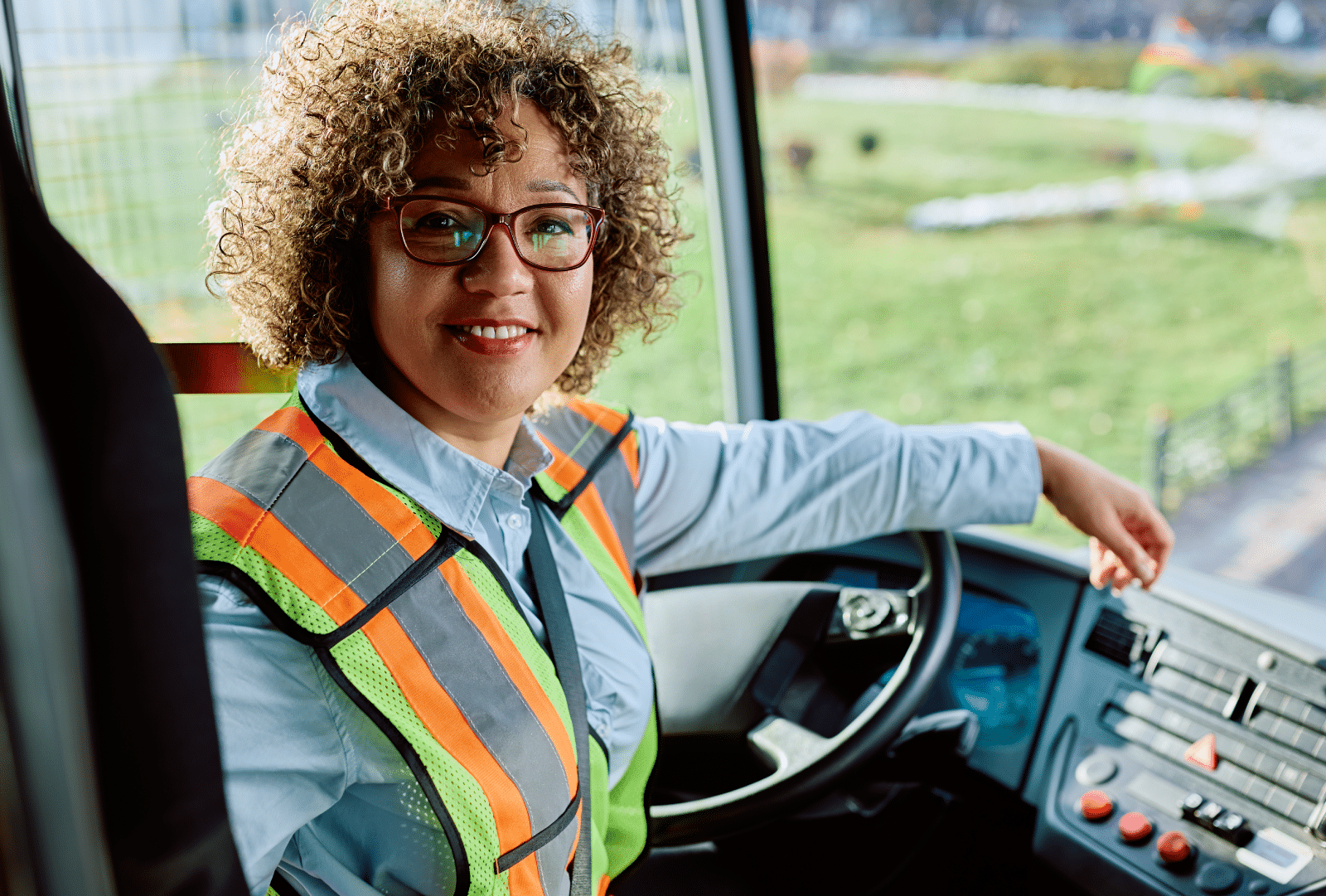 A smiling person with curly hair and glasses, dressed in a safety vest, sits in the driver’s seat of a vehicle holding the steering wheel, and a park can be seen outside the window.