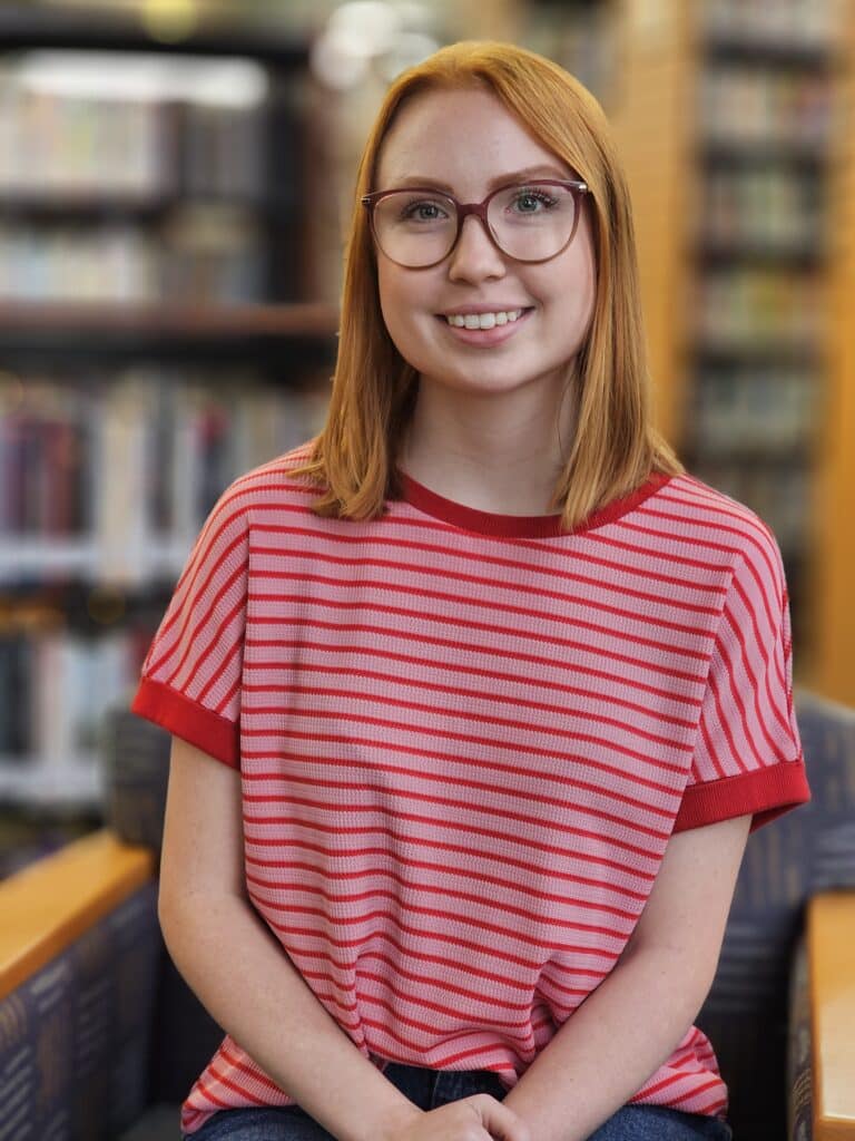 A young woman with red hair and glasses smiles, sitting indoors on a chair. She wears a red and pink striped shirt. The background shows blurred bookshelves, suggesting she is in a library or bookstore.