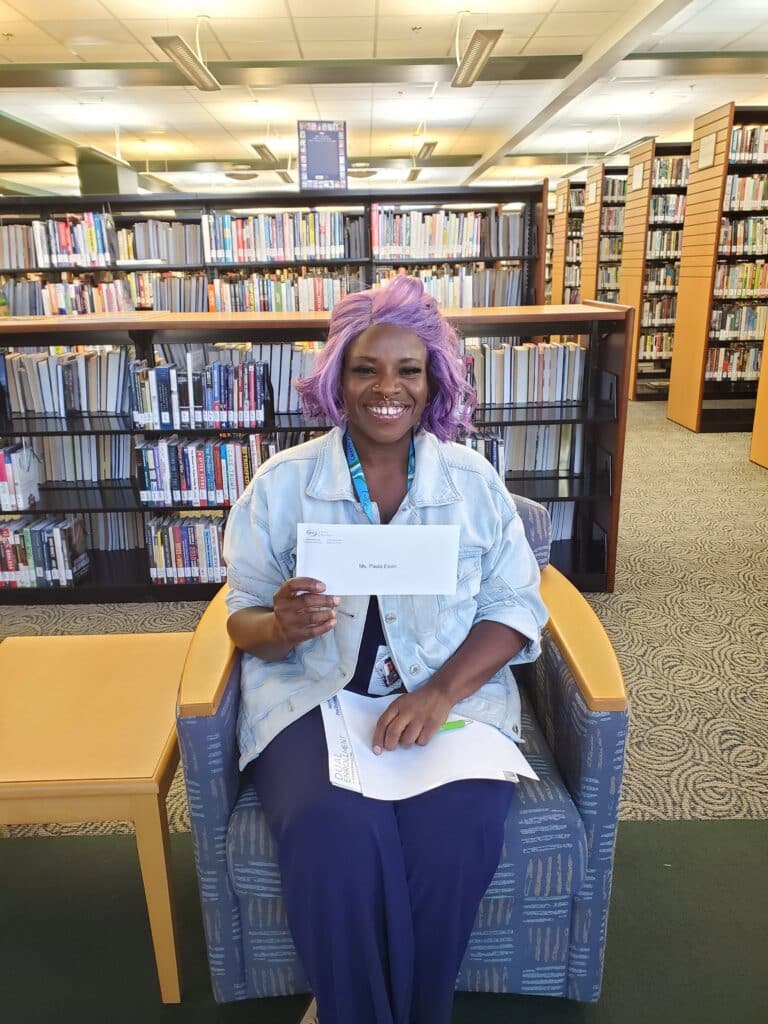 A smiling person with purple hair sits in a library holding an envelope, surrounded by bookshelves. A table with papers and a pen is next to them.