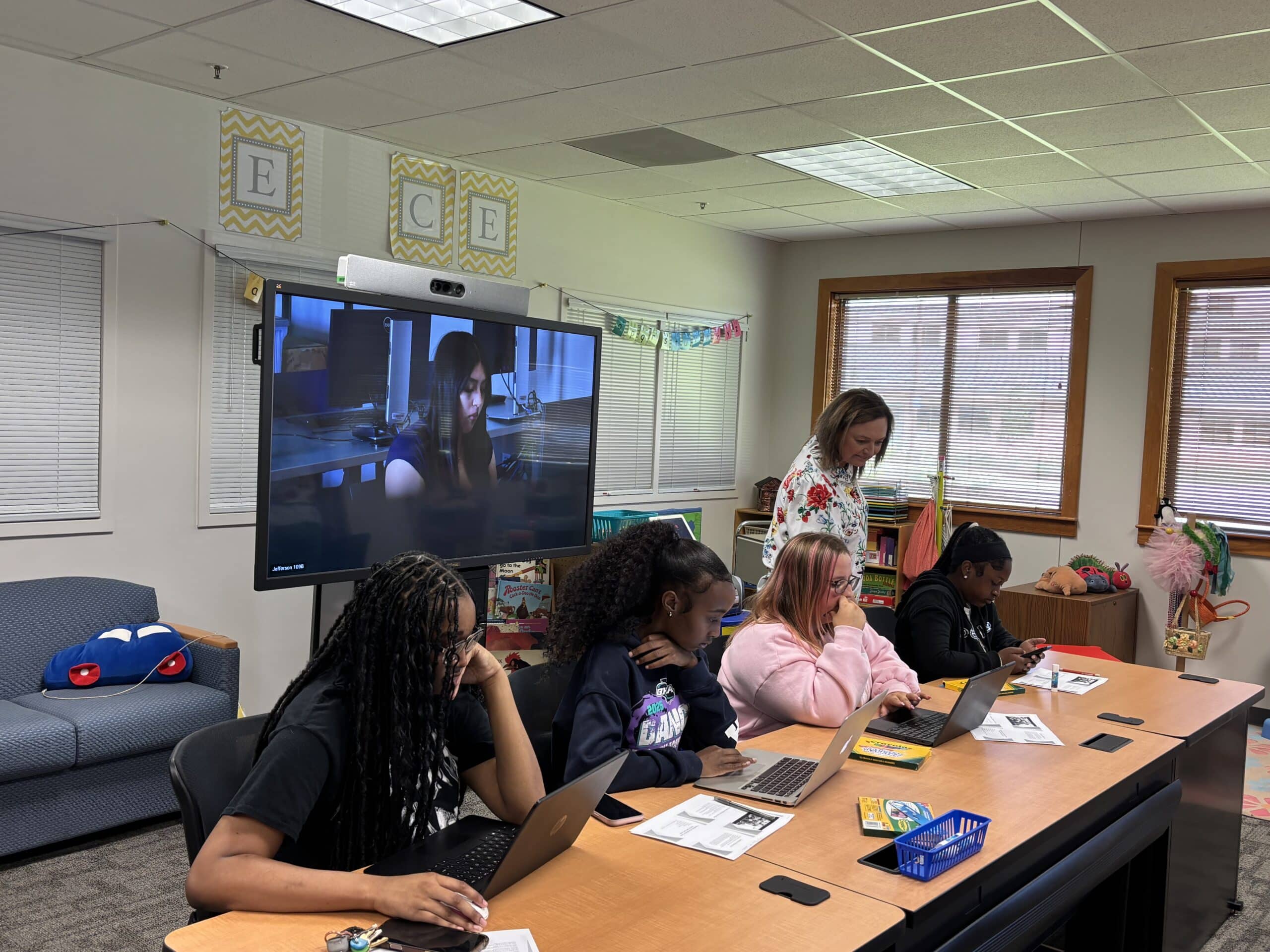 Four OFTC Early Childhood dual enrollment students work at a table with laptops and papers, while a teacher stands behind them. A woman is visible on a large screen at the front of the classroom. The room features windows and bright decor.