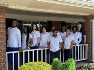Ten people wearing white uniforms pose and smile together on a porch with a white railing, standing in front of a brick building with shrubs in the foreground.