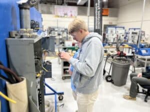 A young man in safety glasses and a gray hoodie works with electrical wiring in a technical lab filled with equipment and machinery.