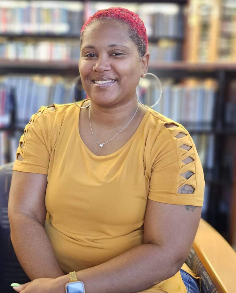 Smiling person with short red hair, hoop earrings, and a mustard-yellow top sits in a library with blurred bookshelves in the background.