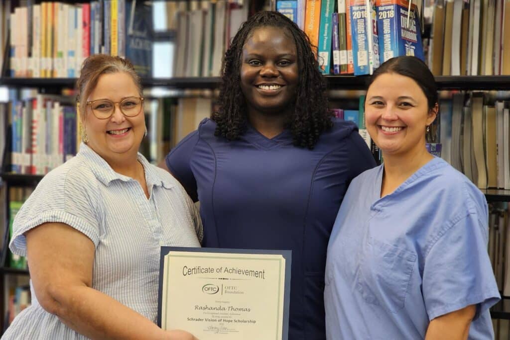 Three women stand smiling in front of bookshelves. The woman in the center, dressed in navy scrubs, holds a framed Certificate of Achievement. The two women on either side wear light-colored tops.