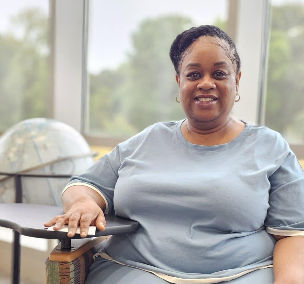 A woman wearing a light blue outfit sits smiling in a chair beside a small desk, with a globe on the desk and large windows behind her revealing greenery outside.