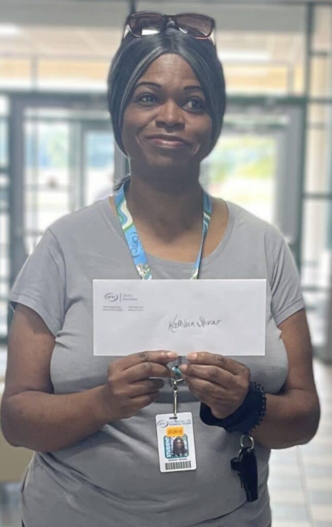 A woman in a gray shirt, wearing a lanyard and badge, smiles while holding an envelope labeled Kathleen Skinner inside a bright indoor space.