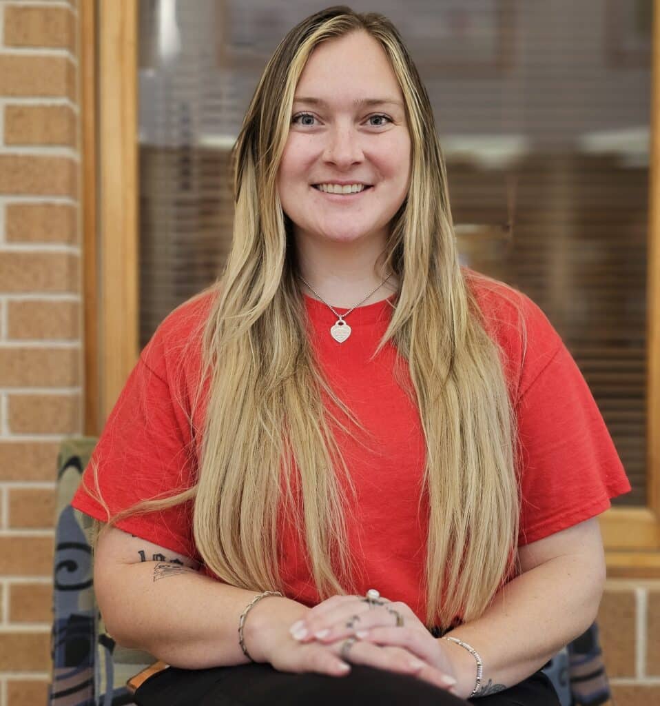 A woman with long blonde hair wearing a red shirt sits smiling with her hands folded on her lap in front of a window and brick wall background.