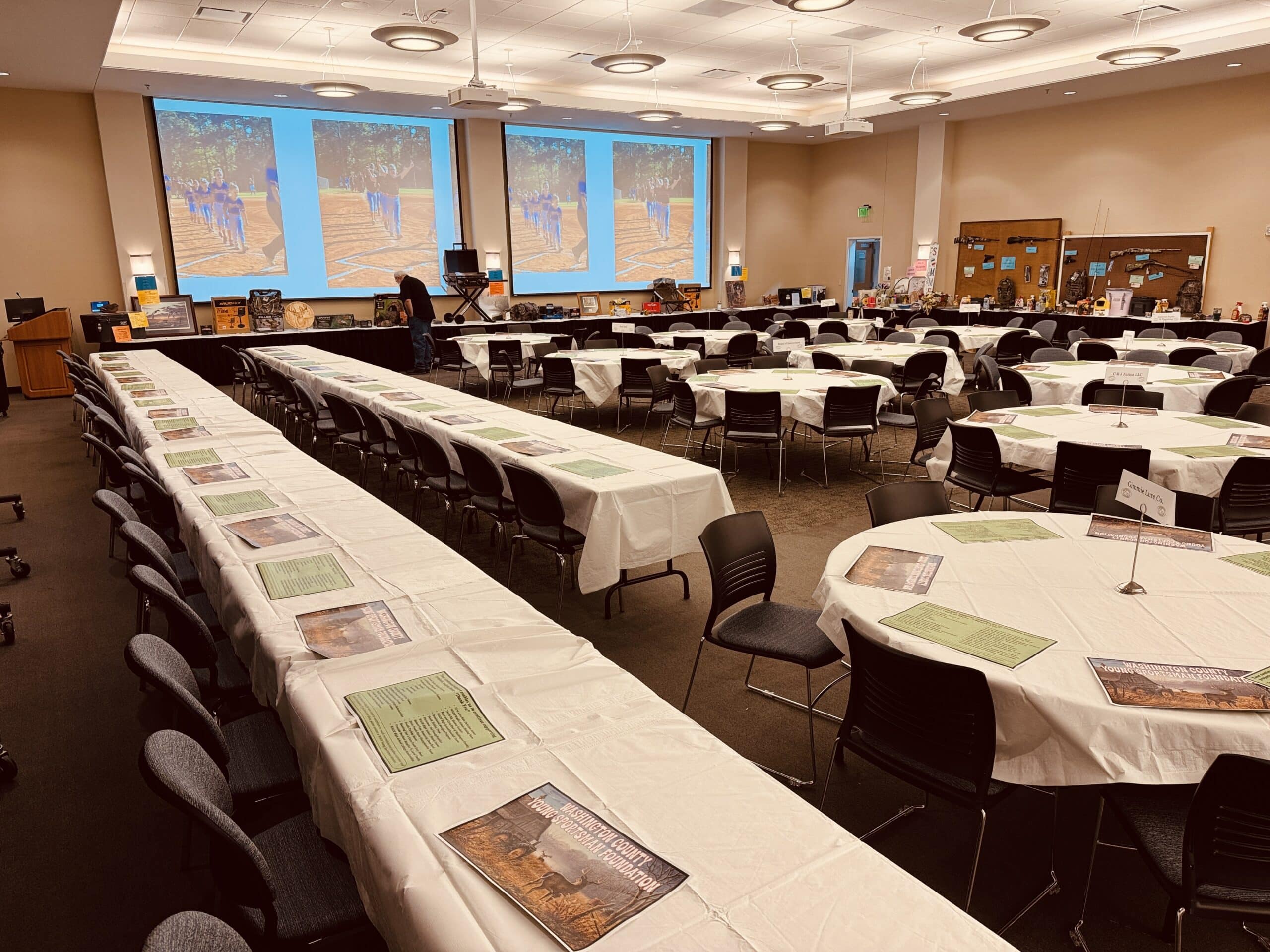 andersville's Tarbutton Center arranged with rows of long tables and circular tables covered in white cloths, with documents placed at each seat and large projection screens at the front displaying photos of people on a track.
