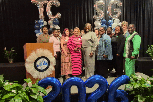 A group of people standing behind a wooden podium at an event, with large silver balloons spelling TC SG and large blue balloons spelling 2026.