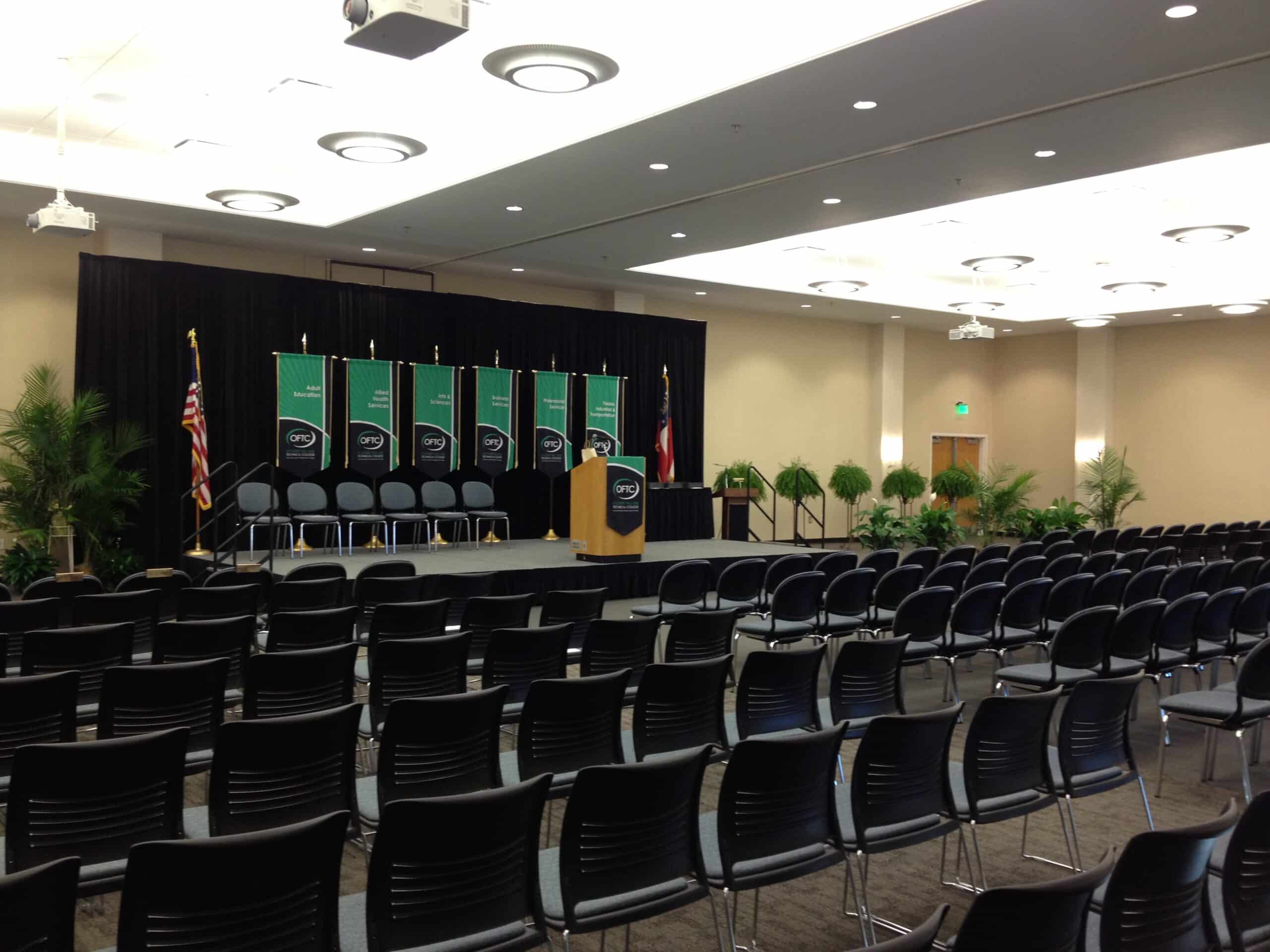Sandersville's Tarbutton Center setup for event with rows of black chairs facing a stage with a podium and several department banners hanging against a black curtain.
